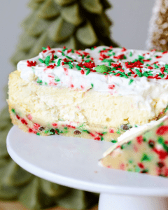 sliced frosted sugar cookie cheesecake with a sugar cookie crust, whipped cream and sprinkles