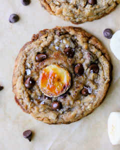 Two, freshly baked banana bread chocolate chip cookies on a baking sheet with bruleed banana slices and flaky sea salt on top.