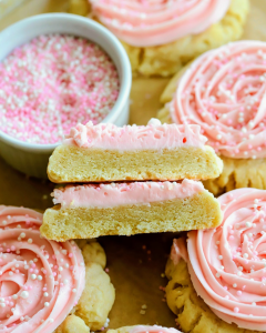 Bakery-style sugar cookies with smooth pink frosting and colorful sprinkles, with one cookie sliced open to show the soft, thick interior.