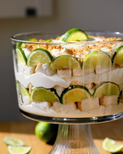 Layered key lime trifle in a glass dish featuring cubed angel food cake, creamy key lime filling, Biscoff and macadamia nut crumble, white chocolate pieces, and a whipped cream topping garnished with lime zest and wedges.
