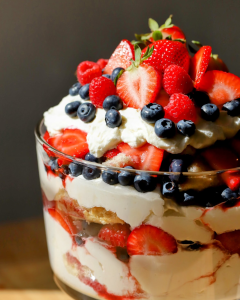 A glass trifle dish filled with layers of vanilla cake, fresh strawberries, raspberries, blueberries, and fluffy mascarpone cream cheese frosting, showcasing each colorful layer of the assembled berry trifle.
