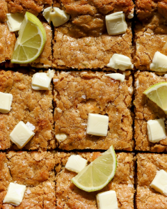 Square slices of lime and toasted coconut blondies made with brown butter, white chocolate, and macadamia nuts, decorated with white chocolate drizzle and fresh lime slices.