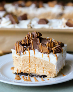 A plated sliced of peanut butter slab pie with a chocolate sandwich cookie crust, thick peanut butter mousse filling, fluffy whipped cream topping, chopped peanut butter cups, and a drizzle of melted peanut butter, sliced and ready to serve.