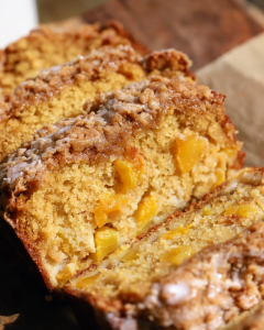 A sliced peach loaf topped with a buttery brown sugar crumble sits in warm afternoon sunlight, showcasing its moist texture and juicy peach pieces. The golden loaf is arranged on a serving board, highlighting the tender crumb and crisp streusel topping.