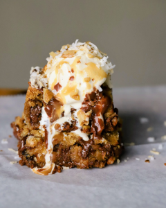 A warm slice of Chili's Copycat Paradise Pie featuring a buttery Biscoff cookie crust and chewy brown butter blondies loaded with shredded coconut, chocolate chips, and walnuts. The dessert is topped with a scoop of vanilla ice cream, drizzled with rich chocolate sauce and salted caramel sauce for the ultimate indulgent copycat treat.