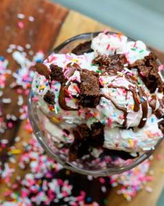 Scoops of homemade cake batter brownie ice cream in a glass bowl topped with colorful sprinkles and a drizzle of chocolate sauce, showing the creamy ice cream with brownie pieces throughout.