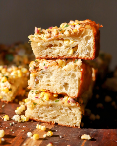 Thick slices of Mexican street corn focaccia stacked with roasted corn, creamy elote sauce, crumbled cotija cheese, fresh cilantro, chili powder, and lime, showcasing the soft, airy crumb of the homemade focaccia.