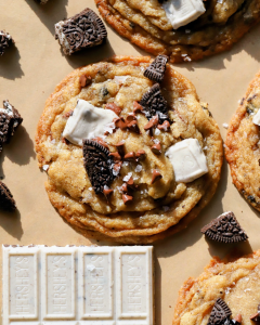 Thin and crispy cookies on a baking sheet, surrounded by chocolate sandwich cookies, cookies and cream candy bars, and sprinkled with flaky sea salt.