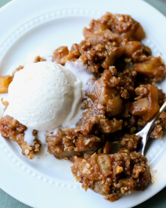 A golden-brown slice of crunchy apple crisp on a white plate, topped with a scoop of creamy vanilla ice cream as it begins to melt over the warm, buttery oat topping.