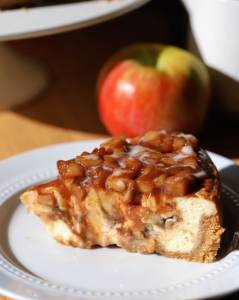 A slice of apple cinnamon roll cheesecake on a white plate, showing golden swirled layers of cinnamon brown sugar, spiced apples, and creamy cheesecake.