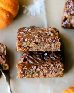 Two sliced pumpkin toffee cookie bars on parchment paper, surrounded by small pumpkins, showcasing the soft, chewy layers and maple toffee topping.