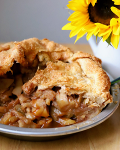A flaky slice of double-crust salted caramel apple pie lifted from the pan on a rustic wooden table.